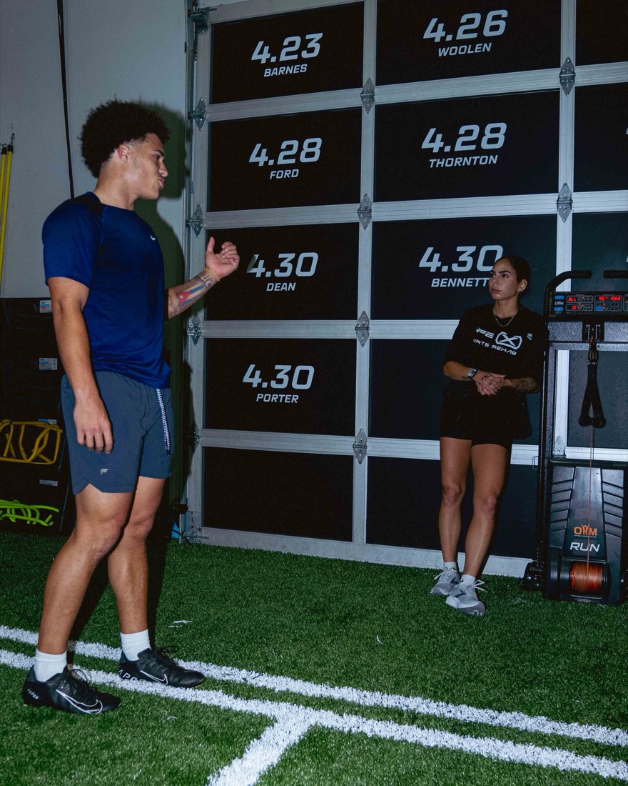 Nicole coaching an athlete on the turf in front of the 40-yard times wall.