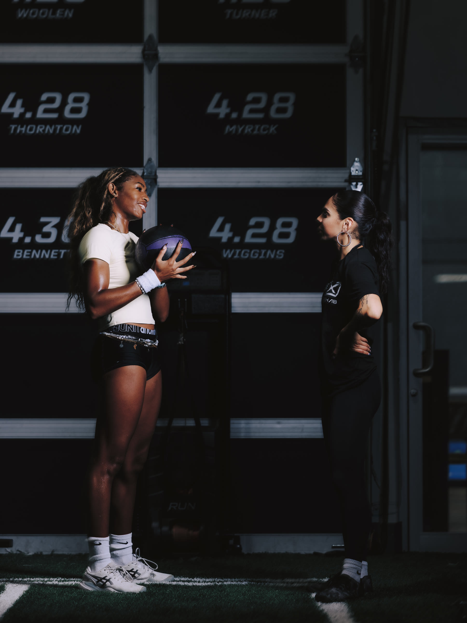 Nicole with an athlete holding a medicine ball in front of the 40-yard times wall.